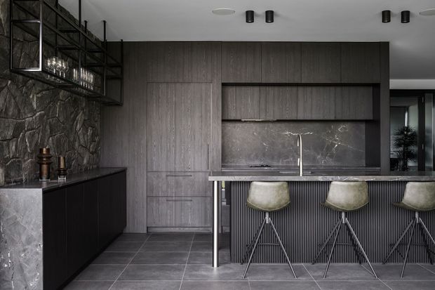 A dark, sophisticated contemporary kitchen and home bar area with floor-to-ceiling timber-look cabinetry, a marble or natural-stone benchtop and backsplash, a vertically ribbed island front, and three bar stools. The room features a dramatic rough-stone feature wall, dark tiled flooring, black metal open shelving with glassware, and recessed and surface-mounted black lighting.