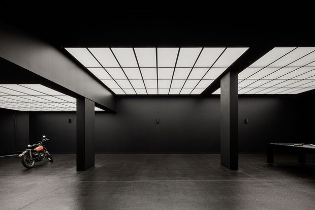 A dark, minimalist interior with black walls, structural square columns, polished dark concrete flooring, and large luminous gridded ceiling panels. A motorcycle is displayed on the left and a pool table or similar display table is partially visible on the right, suggesting a gallery, showroom, or entertainment space.