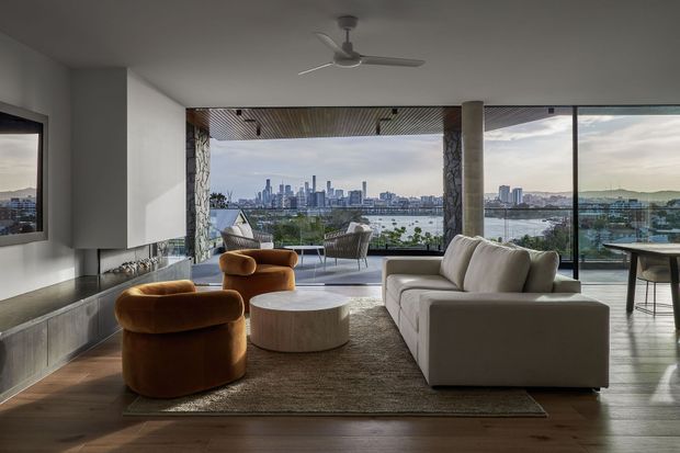A contemporary living room with expansive floor-to-ceiling glazing overlooking a waterfront city skyline. The room features a white sectional sofa, rounded rust-coloured lounge chairs, a low round coffee table, a large neutral rug, dark timber flooring, a wall-mounted television, and a central ceiling fan. A covered outdoor terrace with additional seating is visible beyond the glazing.