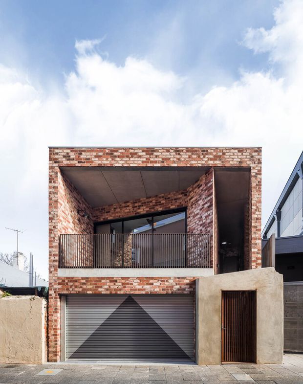 Street-facing view of a compact contemporary multi-storey residence with a red and cream exposed-brick facade. The upper level features a deeply recessed balcony with dark metal balustrading, brick side walls, and a dark soffit. At ground level are a wide grey roller garage door with geometric detailing and a narrow timber entry door set into a textured light render wall. Adjacent modern residential construction is visible at the right.