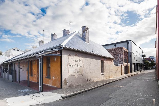 Street-level exterior view of a historic low-rise corner building with weathered rendered walls, a corrugated metal hipped roof, multiple brick chimneys, verandah-style covered frontage, and heritage signage reading “Fremantle Angel’s House”. A contemporary multi-storey building adjoins it along the narrow urban street.