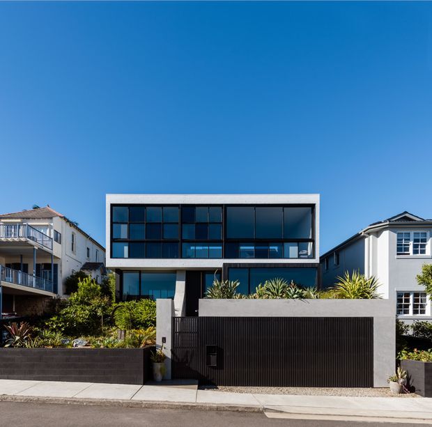 Street-facing photograph of a contemporary multi-storey residence with a strong rectangular form, broad dark-framed glazing, white perimeter framing, and a dark vertically ribbed garage door. Layered raised garden beds and tropical-style planting sit behind a low dark masonry wall, with neighbouring residential buildings on either side under a clear blue sky.