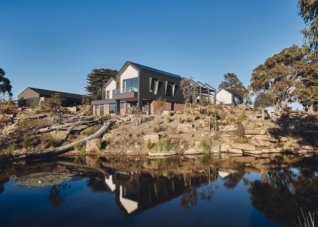 Exterior view of a contemporary multi-level house set on a rocky, landscaped site beside a reflective pond. The home has dark vertical cladding, pale rendered gable walls, large glazed openings, projecting upper-level forms and a partially open pergola or terrace structure. Additional residential structures and mature trees surround the property.