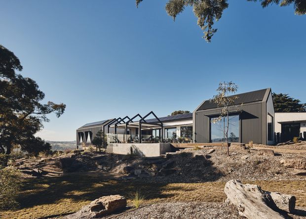 Exterior view of a contemporary single-storey home set on a rocky, sloping landscape. The building has dark grey vertical cladding, pitched gabled roof forms, large glazed openings, and a partially enclosed terrace or veranda with repeated exposed gable-like structural frames. Native trees and distant rolling terrain surround the house.