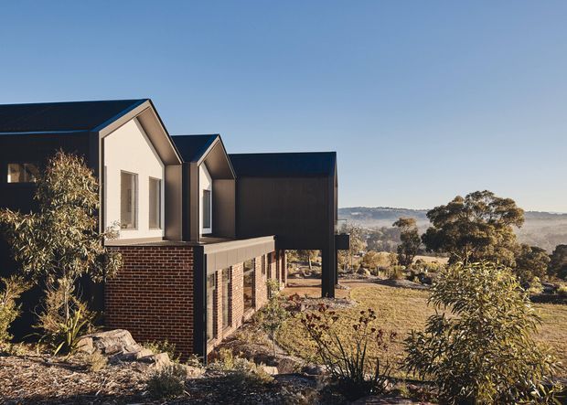 Exterior view of a contemporary multi-level residential building on a dry, sloping landscape. The facade combines dark metal cladding, pale gabled wall sections, exposed red brickwork, and extensive glazing, with projecting upper volumes and a strong cantilevered form. Native-looking vegetation and a broad rural or semi-rural valley landscape surround the house.