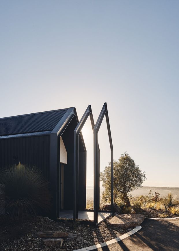 A compact dark-clad building set on a landscaped hillside, featuring a sharply angular pitched roof and a projecting skeletal veranda or entry frame. The image is photographed from outside in warm low sunlight, with native grasses, rocks, a tree, and a broad distant landscape visible beyond.