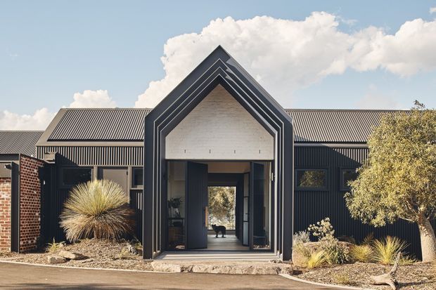 Front exterior view of a contemporary single-storey building with a dramatic nested gable entrance canopy, dark vertical metal cladding, corrugated pitched roofing, exposed brick side walls, and a glazed entry opening. Drought-tolerant landscaping with ornamental grasses and shrubs frames the facade.