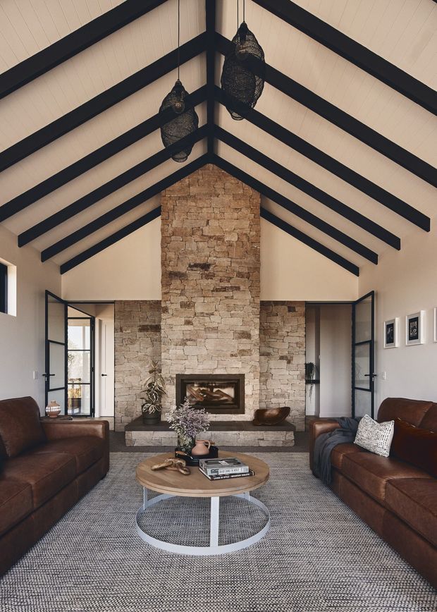 A refined living room with a dramatic vaulted ceiling supported by exposed dark timber beams and rafters. A full-height natural stone fireplace forms the focal point, with black-framed glazed doors opening to adjacent spaces. Two deep brown leather sofas face a round timber-and-metal coffee table over a textured neutral rug. Decorative woven pendant lights, framed artwork, and understated furnishings create a warm, rustic-contemporary atmosphere.