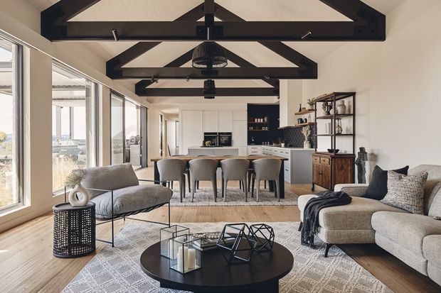 Open-plan living and dining area with a kitchen at the rear, featuring exposed dark timber roof trusses, white paneled ceilings, extensive glazing, neutral upholstered seating, a round black coffee table, timber flooring, and open shelving with decorative storage. The room has a bright, refined contemporary-rustic character and appears spacious with a high pitched ceiling.