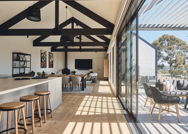 A bright, open-plan contemporary interior combining a kitchen, dining area and living space. A white island with timber stools faces a dining table and lounge area, while exposed dark timber roof trusses and black pendant lights create strong architectural contrast. Full-height glazed sliding doors open to a furnished outdoor terrace, allowing abundant sunlight and views of the surrounding landscape.