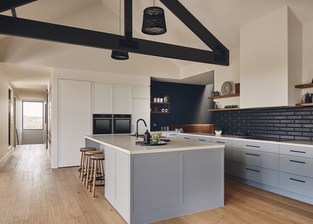 A bright, open-plan kitchen with a large central island, integrated double ovens, white cabinetry, pale stone benchtops, and a black glazed brick splashback. The kitchen opens to a hallway and features exposed dark timber roof trusses, a pitched ceiling, timber flooring, open timber shelving, and pendant lighting. The palette is predominantly white, light wood, charcoal, and deep navy.