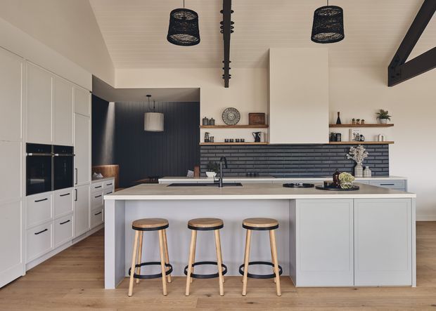 A spacious contemporary kitchen with a large light-coloured island and three timber-and-black stools, white full-height cabinetry with integrated appliances, a dark horizontal brick-tile feature wall, open timber shelving, and warm timber flooring. Black pendant lights and exposed dark structural beams add contrast, while a dark-lined opening leads toward an adjoining space.