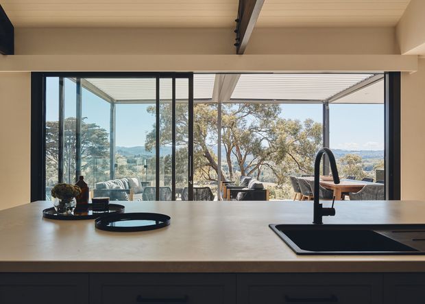 A contemporary kitchen viewed from behind a broad island benchtop, with dark lower cabinetry, a deep dark sink and black gooseneck tap. Large black-framed sliding glass doors open the kitchen toward a bright outdoor terrace and scenic tree-covered landscape. The room has a warm timber-lined ceiling with exposed structural beams and strong indoor-outdoor connection.