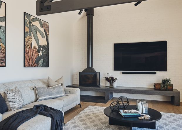 A warm contemporary living room with a light upholstered sectional sofa, patterned area rug, round black coffee table, and a long dark built-in media console. A wall-mounted television is set against a white-painted brick wall, with a compact black fireplace and exposed flue beneath a substantial dark overhead structural beam. Botanical artwork and potted plants add natural decorative accents.