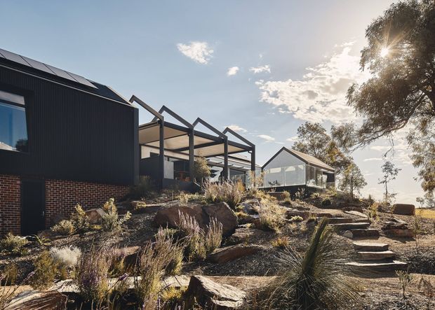 A contemporary hillside residential exterior set within a dry, natural landscape. The building combines dark corrugated cladding and exposed red brick with a glazed, open-framed pergola or connecting structure and a smaller gabled pavilion. Stone steps and rock retaining elements lead through drought-tolerant planting toward the house. Strong afternoon sunlight and mature trees frame the scene.