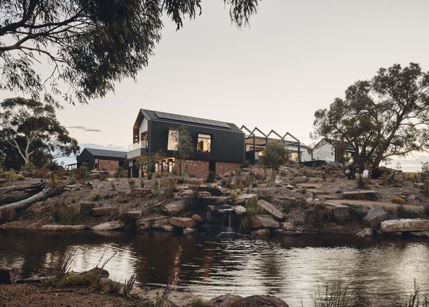 Exterior view of a contemporary multi-level dwelling set within a rocky, landscaped site beside a pond or water feature. The building combines dark metal-clad upper volumes with exposed red-brown brick lower walls, pitched roof forms, extensive glazing, and a partially visible open-frame terrace or pavilion. Mature eucalyptus-like trees, natural stone landscaping, and reflective water create a rugged Australian bush setting.