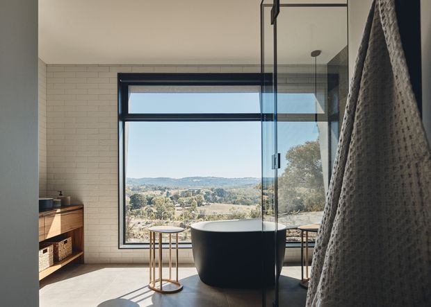 A refined contemporary bathroom viewed through a frameless glass shower screen, featuring a freestanding matte-black oval bathtub, light brick-pattern wall tiling, a large black-framed picture window, timber vanity or storage joinery, and a scenic leafy hillside outlook. A textured neutral towel or robe hangs in the foreground.