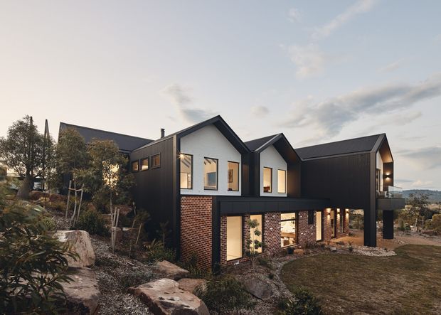Exterior architectural photograph of a contemporary multi-level house set on a rocky, sloping site. The home features multiple intersecting pitched gable roofs, dark vertical cladding, white upper-level walls, exposed red brick at the lower level, large windows, and a projecting upper volume supported by dark columns. Landscaping and distant residential surroundings are visible.