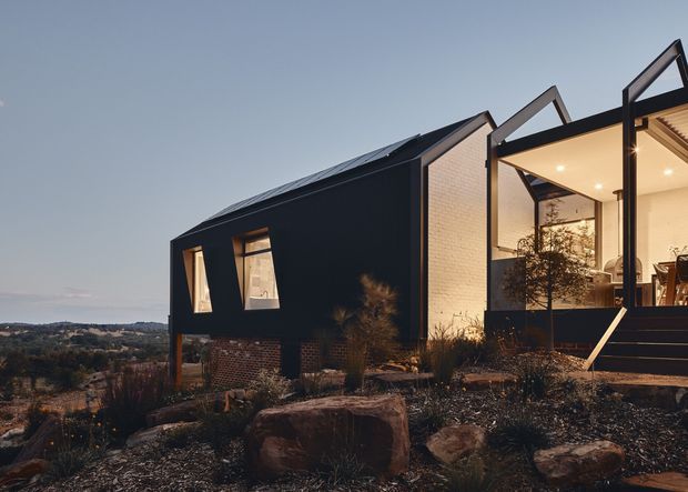 A contemporary angular house viewed from the exterior at dusk, set on a rocky, landscaped sloping site. The dark-clad main volume has a steep pitched roof with visible solar panels, narrow irregular windows, and a contrasting brick base. An open glazed entry or living area projects at right, with warm interior lighting, timber-lined soffits, and an external stair. The distant rolling landscape is visible beyond the property.