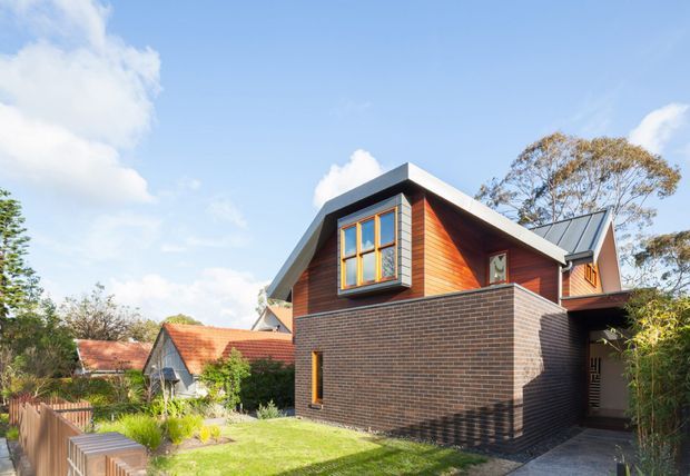 Exterior view of a contemporary two-storey house extension or upper-level dwelling, featuring dark brickwork, warm timber cladding, a projecting timber-framed window, and a steeply pitched metal roof. The home sits in a leafy suburban garden setting with neighbouring pitched-roof homes visible in the background.