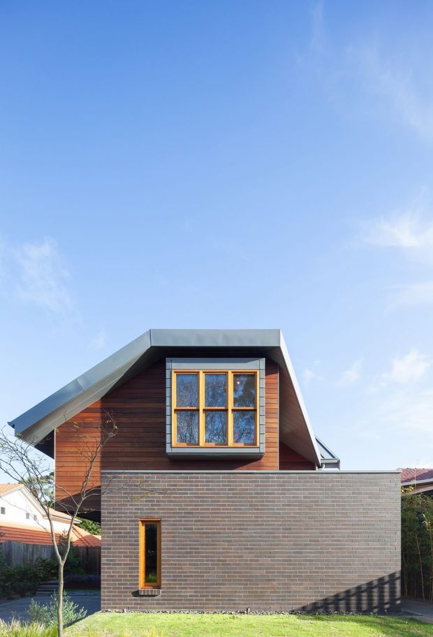 Exterior view of a compact contemporary residential extension or house, featuring a steep asymmetrical gabled roof with broad metal edging, dark horizontal timber cladding to the upper storey, and a lower facade of grey-brown brickwork. A projecting multi-pane timber-framed window forms a prominent upper-level feature, with a narrow vertical window below. The image is photographed from the garden under a clear blue sky.