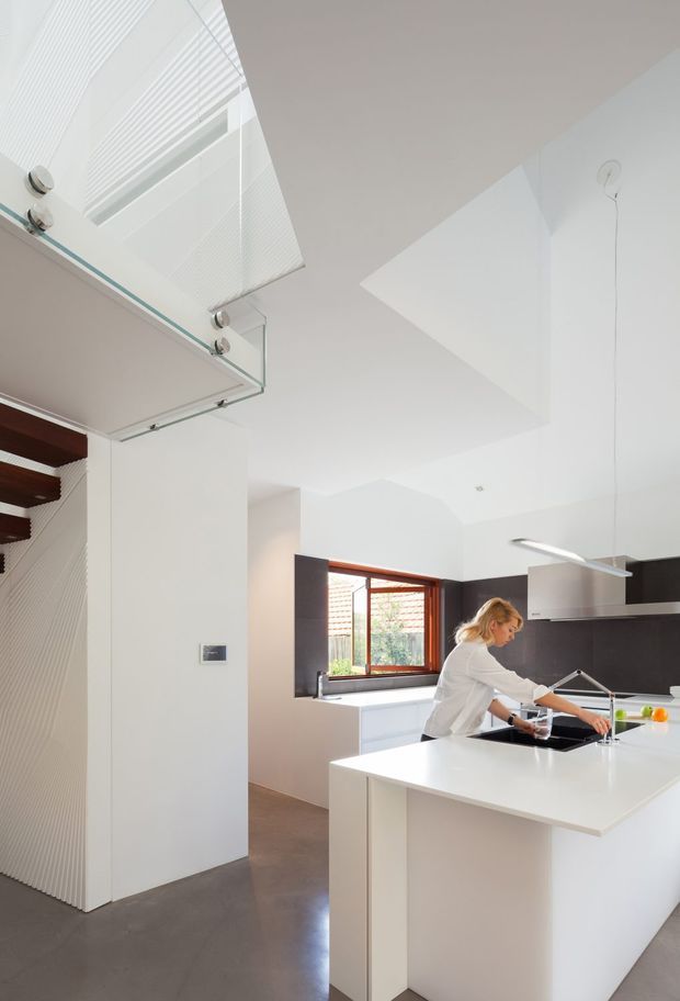 Bright contemporary kitchen interior with a large white island and integrated sink, dark cabinetry and splashback, white walls, and a timber-framed window above the rear bench. The open-plan space includes a sculptural staircase and a glass balustrade leading to an upper level. A person is working at the island.