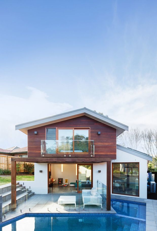 Exterior view of a contemporary two-storey home with a prominent gable roof, warm horizontal timber cladding on the upper level, white rendered walls below, extensive glazing, glass balcony balustrades, and a covered outdoor living area opening toward a swimming pool. The house sits in a landscaped suburban setting and is photographed in daylight.