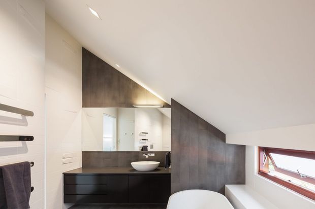 Contemporary bathroom beneath a sloping ceiling, featuring dark timber-toned wall panelling and vanity cabinetry, a white vessel basin, mirrored wall, recessed lighting, and a partially visible freestanding bathtub. A high horizontal window provides natural light.