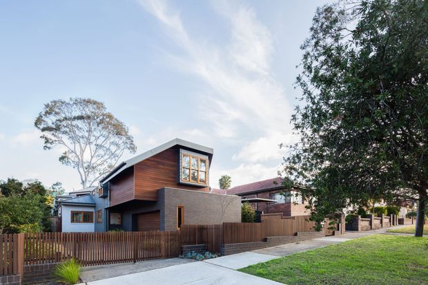 Exterior streetscape view of a contemporary multi-level residential home with dark horizontal cladding, warm timber wall panels, a projecting upper volume, large windows, and a low-pitched roof. The house is set behind timber fencing with a paved driveway and landscaped suburban frontage; neighboring homes are visible along the street.