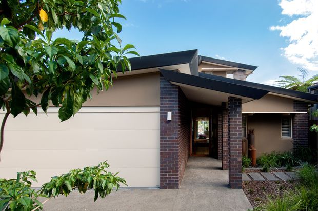 Contemporary two-storey residential house viewed from the front, featuring a broad double garage, dark angular metal roof planes, a projecting entry canopy supported by dark brick columns, rendered walls, and a covered walkway leading into the home. The front garden includes low planting and concrete stepping slabs, with a leafy tree partially framing the view.