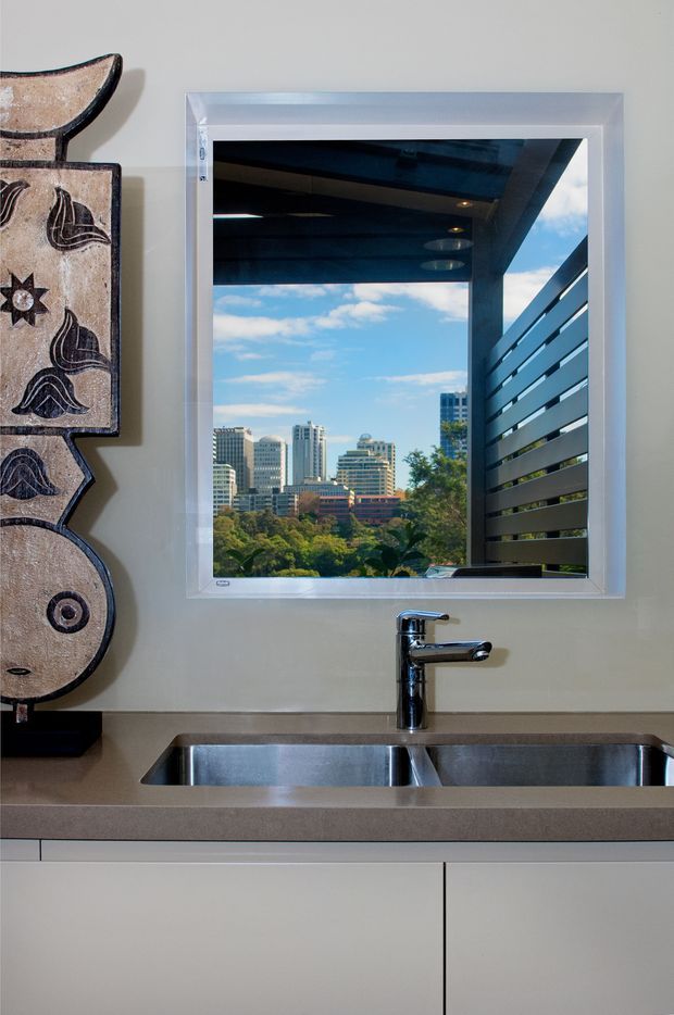 Close-up view of a compact contemporary kitchen sink area beneath a framed window. The space features a stainless-steel double-bowl sink, chrome mixer tap, taupe stone-look benchtop, and smooth white lower cabinetry. Through the window is a covered outdoor area with horizontal screening and a leafy urban skyline beyond. A decorative carved wall artwork is visible at the left edge.