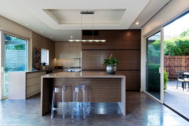 Contemporary kitchen with a large central island, integrated cabinetry, dark horizontal timber wall panelling, polished concrete-look flooring, and extensive glazing opening to an outdoor patio. Transparent bar stools and pendant lighting sit beneath a recessed ceiling feature.