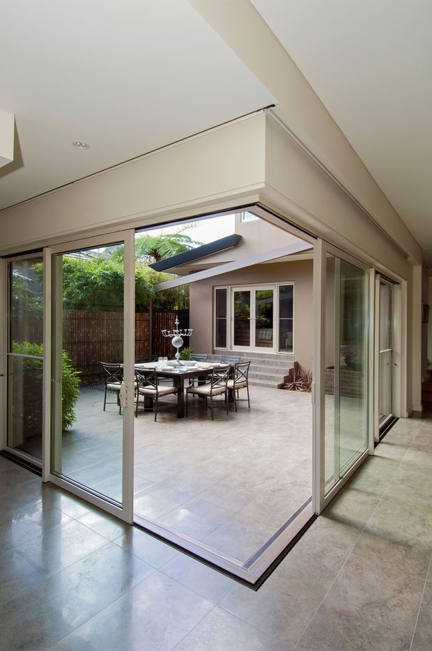 Interior view looking through wide-open glazed folding or sliding doors onto a landscaped courtyard and outdoor dining area. The room has pale tiled flooring, white walls and ceiling, extensive glass framing, and a covered patio beyond with a dining table and chairs. The image emphasizes the strong indoor–outdoor connection and courtyard outlook.