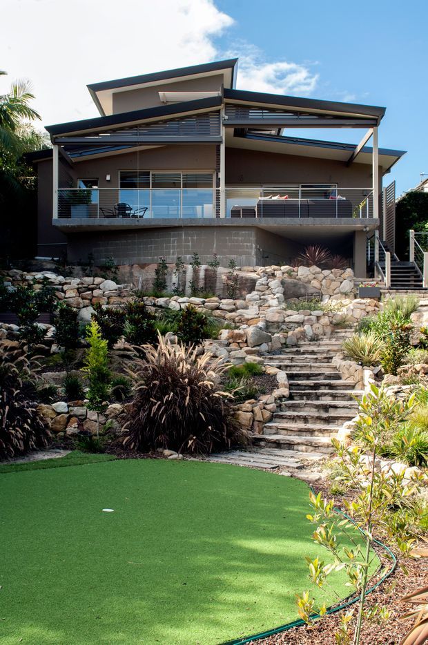 Exterior view of a contemporary multi-level house on a landscaped sloping site. The home features angular skillion-style roof forms, broad covered balconies, dark metal roof edges, horizontal louvres, glass balustrades, and rendered walls. A stone retaining landscape with stepped pathways and planted garden beds descends toward a green artificial-turf recreational area in the foreground.