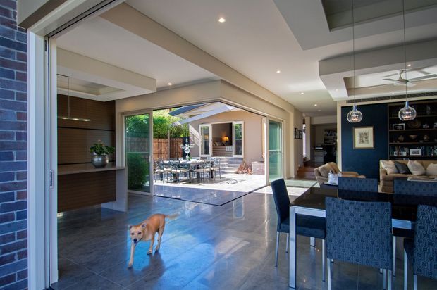 Open-plan contemporary residential interior viewed through large sliding glass doors, combining a dining area, kitchen, and living room around a bright central courtyard. The space has polished dark stone or tile flooring, white recessed ceilings, extensive glazing, dark timber kitchen cabinetry, and a deep blue feature wall with built-in shelving. A covered outdoor dining courtyard is visible beyond the open doors.