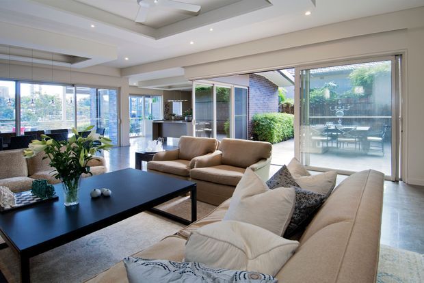 Bright, spacious contemporary open-plan living room with large beige sofas, a black rectangular coffee table, pale flooring, and extensive sliding glass doors opening to a landscaped courtyard or patio. Recessed ceiling lighting, a ceiling fan, white built-in ceiling detailing, and abundant glazing create an airy indoor-outdoor connection. A kitchen and dining area are visible in the background.