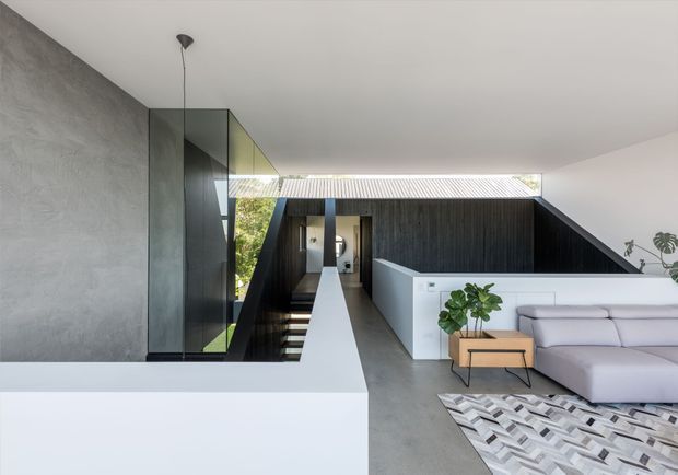 A minimalist contemporary upper-level living area with a pale grey polished concrete floor, white built-in half-height walls and cabinetry, a large light-grey sectional sofa, and a black vertically ribbed feature wall. A glass balustrade overlooks a stairwell, while a linear skylight or clerestory brings daylight into the space. The restrained monochrome palette is softened by indoor plants and a geometric monochrome rug.