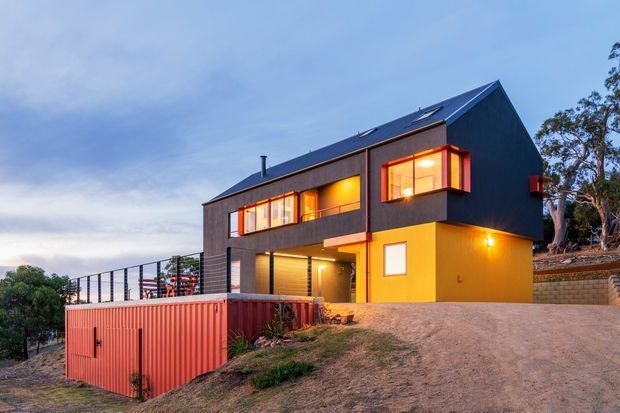Exterior view of a contemporary two-storey house on a sloping site at dusk. The building has dark vertical cladding, bold warm yellow and red/orange accent walls, angular roof forms, projecting bay windows, roof skylights, and a red metal garage or storage structure integrated into the lower foreground. A terrace with slender black railings and surrounding trees are visible.