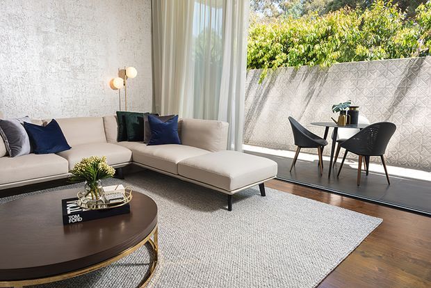 A bright, refined contemporary living room with a large cream sectional sofa, dark round coffee table, textured light rug, and sculptural globe floor lamp. Full-height glazing opens onto a compact outdoor dining area with black chairs and a small round table, screened by a patterned concrete privacy wall. Dark timber flooring and neutral finishes create a warm, polished interior.