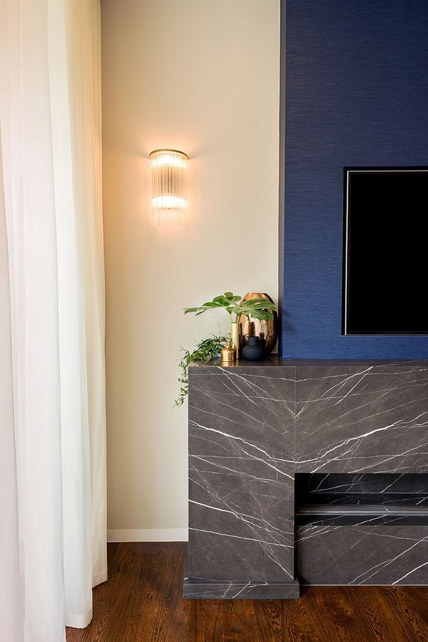 A refined living room media wall featuring a dark blue textured feature wall, wall-mounted television, and a built-in fireplace surround clad in dramatic dark veined stone. Warm timber flooring, sheer full-height curtains, a decorative wall sconce, and potted greenery create a luxurious contemporary setting.