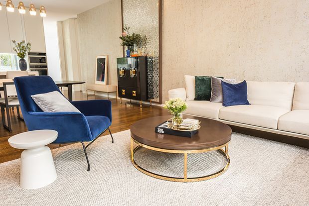 A refined open-plan living room with a white sofa, deep blue upholstered lounge chair, round dark wood coffee table with a brass-toned metal base, white side table, and a large neutral rug. The room features warm timber flooring, textured beige walls, contemporary pendant lighting, and a decorative mirrored and mosaic-panel bar or display joinery unit. A dining area and kitchen are visible in the background.