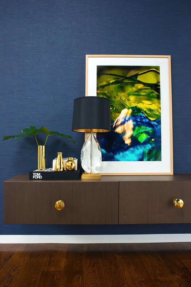 A styled interior vignette featuring a dark navy textured wall, a floating dark wood sideboard with fine vertical grain and round gold knobs, and a framed abstract artwork in vivid green, blue and yellow tones. Decorative objects include a sculptural table lamp with a black shade and faceted clear base, gold-toned vessels, a small plant, and a black book. Dark timber flooring and a white skirting board complete the scene.