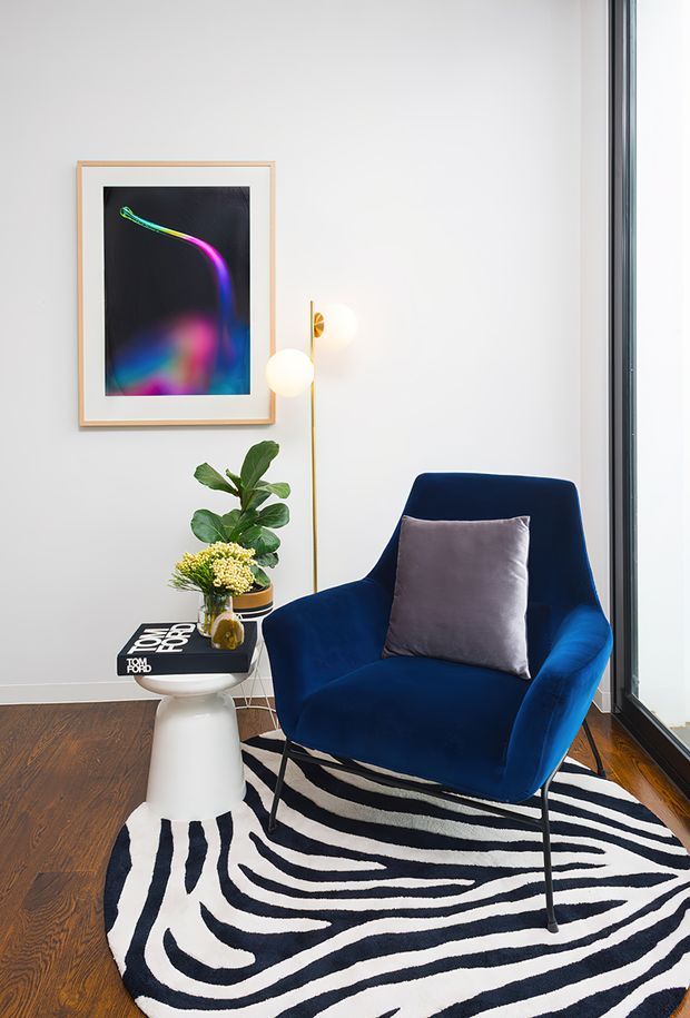 A styled reading nook or seating corner with a deep blue upholstered armchair, grey cushion, white pedestal side table, potted plant, globe floor lamp, framed abstract artwork, and a bold black-and-white zebra-pattern rug. The setting has white walls, dark timber flooring, and full-height black-framed glazing, creating a contemporary, graphic interior.