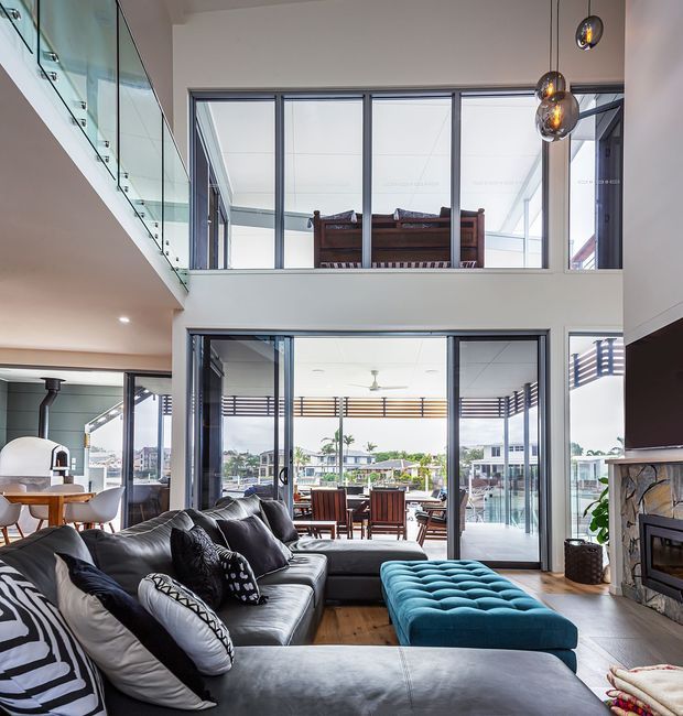 A spacious contemporary double-height living room with a large black leather sectional sofa and bright turquoise tufted ottoman. Full-height glazing opens to a waterfront-facing outdoor dining terrace, while a glass balustrade leads to an upper-level mezzanine. The room includes a stone-clad fireplace, timber flooring, sculptural pendant lights, and expansive windows creating a bright, open atmosphere.