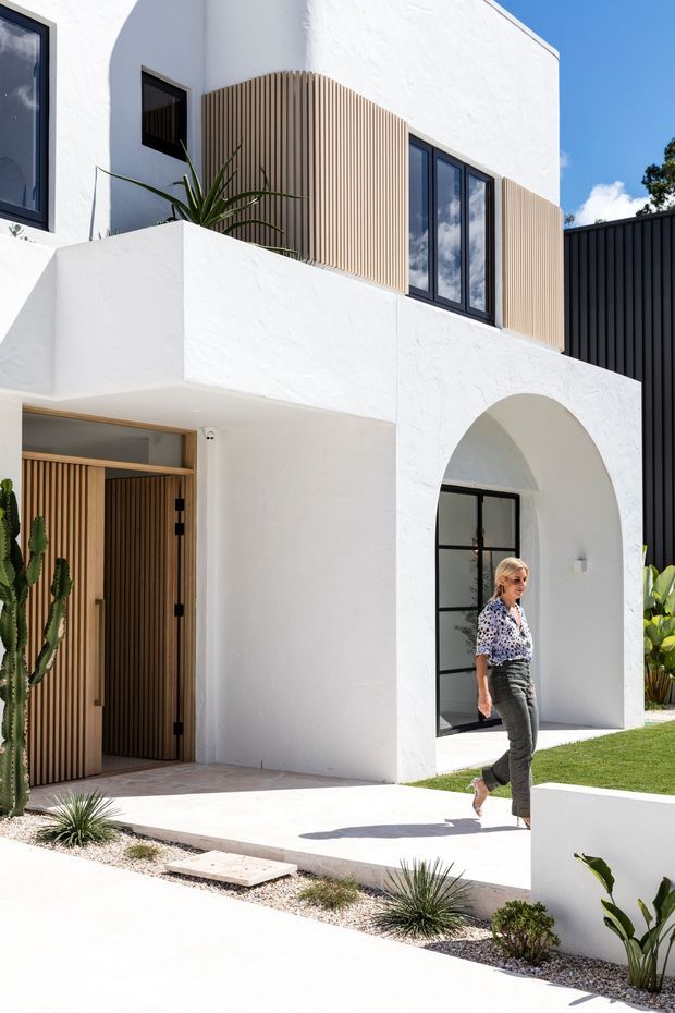 A bright two-storey contemporary home exterior with white textured stucco walls, rounded arches, black-framed windows, vertical timber screening, and a sculptural timber entry door. The facade opens onto a pale paved path with minimalist drought-tolerant landscaping and gravel beds.