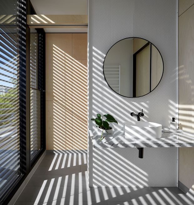 Bright contemporary bathroom with a wall-mounted white vessel basin, black fixtures, round frameless mirror, pale textured wall tiles, and a grey stone vanity benchtop. Strong sunlight filters through external or internal horizontal slats, casting dramatic striped shadows across the walls and floor. Dark-framed glazing is visible on the left, with pale timber-toned built-in cabinetry or doors.