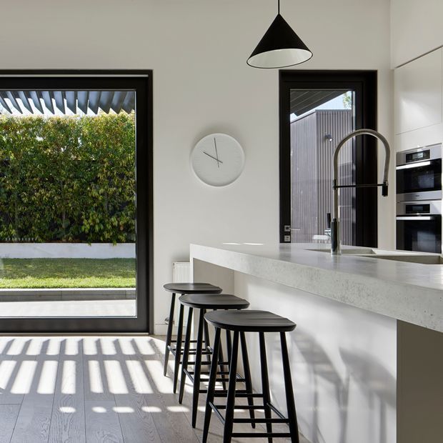 Bright contemporary kitchen featuring a long pale stone island with integrated seating, three black stools, a sculptural black pendant light, dark-framed glazing and door, white cabinetry, built-in ovens, and a statement arched tap. Strong linear sunlight and shadows fall across the pale timber-look floor, with a landscaped garden visible outside.