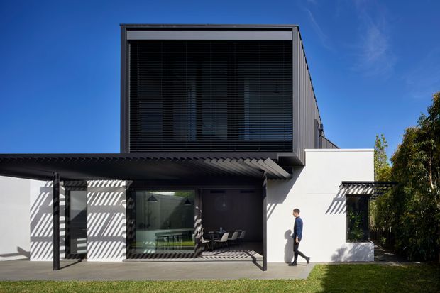 Exterior view of a contemporary two-storey residence with a strong rectangular upper volume, extensive dark louvred glazing, white rendered walls, and a projecting corrugated canopy over the ground-floor terrace. Slim pergola structures cast striped shadows across the facade and paved outdoor area.