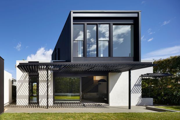 A contemporary two-storey residential facade with a dark upper-level box, expansive black-framed glazing, and a white lower level. A broad slatted pergola projects over the ground-floor outdoor living area, casting strong linear shadows across the walls and paving. The building sits beside a neatly landscaped lawn and hedge under a clear blue sky.