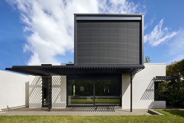 Modern two-storey residential facade photographed from the garden. The building has crisp white rendered walls, dark aluminium-framed glazing, a projecting flat roof or pergola with strong horizontal slatted shading, and a prominent upper-level screened opening with dark horizontal louvres. The geometric shading creates bold striped shadows across the walls and paving, with lawn and planting around the exterior under a bright blue sky.